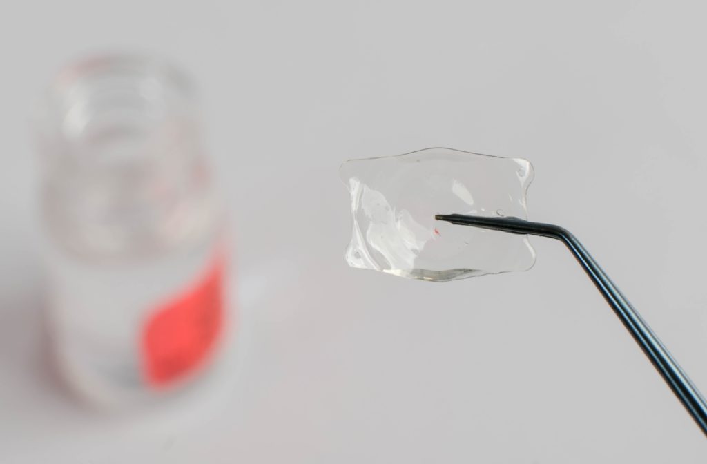 Close up of an Implantable Collamer lends being held up by medical forceps with blurred out bottle of solution in the background.