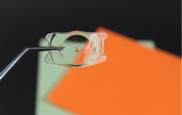 A pair of fine tweezers holding up an implantable collamer lens in front of green and orange pieces of paper