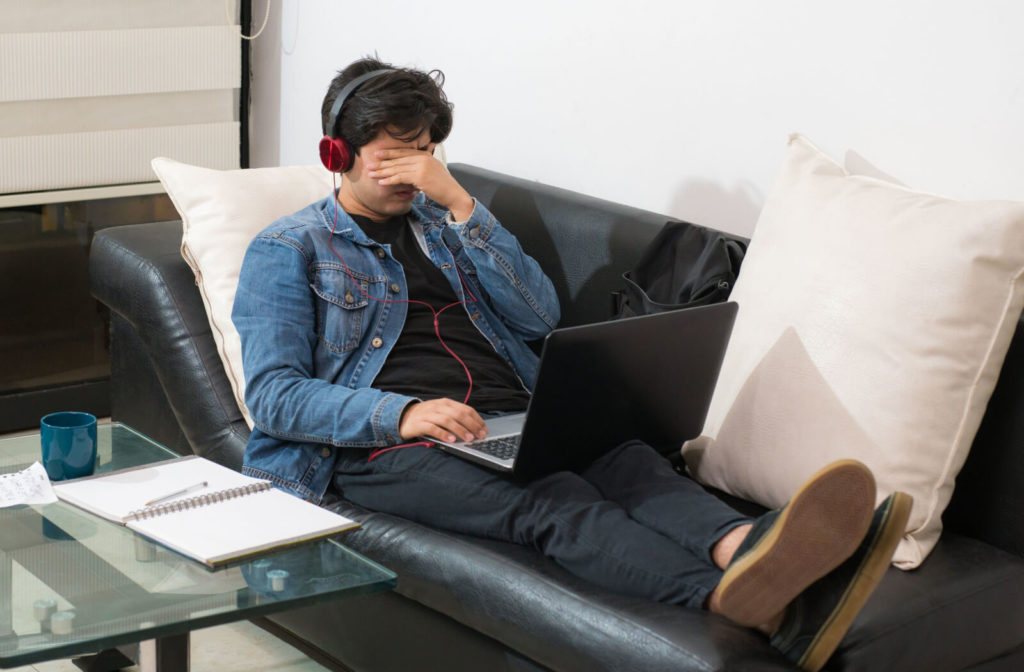 A man sitting on a leather sofa with his laptop on his lap is covering his stressed eye with his left hand after a hours on the screen.