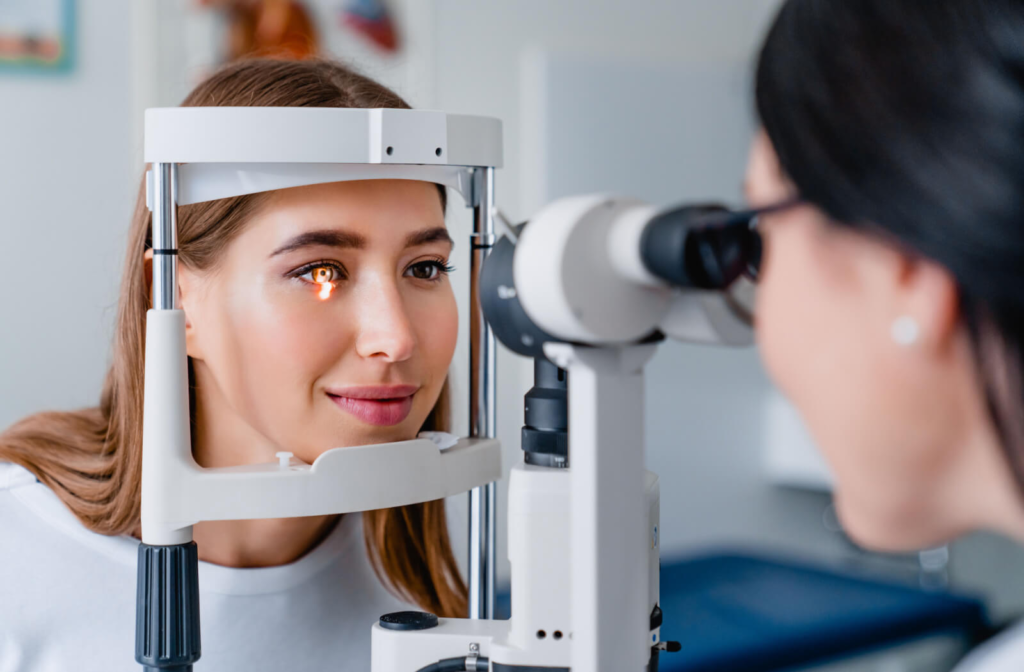 A woman looking into a slit lamp with a light shining in her right eye as her optometrist performs an exam.