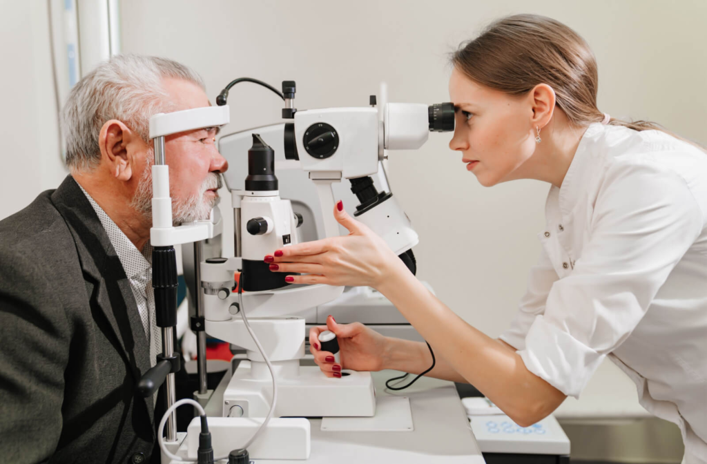 A side view of a senior man looking into a machine while a younger female optometrist examines his eyes.