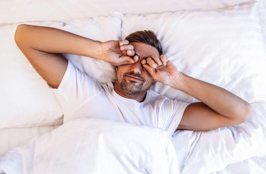 A man lying in bed rubbing his eyes with both of his hands.