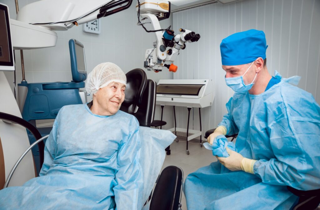 An older adult in blue surgery clothing and a white hairnet is about to lay back on a surgery bed, smiling at a doctor. The doctor is wearing blue scrubs and a blue hairnet, putting on gloves, and laughing with the patient.