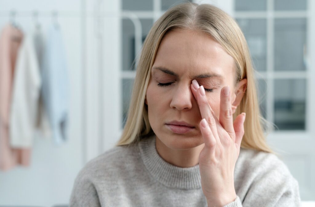 A person in a grey sweater with their eyes closed rubs their left eye in discomfort from meibomian gland dysfunction